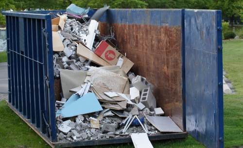 A large blue metal skip positioned outdoors on a grassy area, filled with a mix of construction and household waste including broken concrete, plasterboard, drywall, cardboard boxes, and various discarded materials. The interior of the skip shows rusted metal panels and a wooden plank lining one side, with the waste piled up towards the top, some items spilling over the edges. Surrounding the skip, there is a neatly maintained lawn with trees and greenery in the background, indicating a residential or park setting. The scene is well-lit with natural daylight, emphasizing the textures of the debris and the skip's weathered surface. This image exemplifies general rubbish removal needs that Hornsey House Clearance services could assist with, especially for heavy DIY waste or garden clearance in London postcodes.