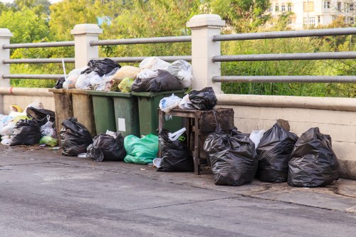 A collection of black plastic rubbish bags, some filled with waste, are piled along the edge of a paved street next to a low concrete barrier with vertical metal railings on top. Several additional bags in green, white, and transparent materials are stacked against the bags. Behind the barrier, there are trees with green foliage and glimpses of multi-storey residential buildings, suggesting an urban or suburban environment. The rubbish bags appear to contain household waste or general refuse, and the scene is outdoors during daylight, with natural lighting. The setting indicates a typical area for rubbish removal or street cleaning, which Hornsey House Clearance services may attend to, especially in residential neighbourhoods around the London postcode area.