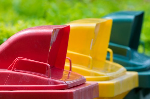 A row of four large plastic rubbish bins positioned on a grassy outdoor area, with the first bin in red, followed by a yellow bin, a green bin, and a blue bin in the background. The bins have textured, slightly reflective surfaces and are fitted with handles and lids. They are arranged in a line, with the red bin in the foreground occupying most of the image, and the other bins partially visible behind it. The setting appears to be a residential or communal garden space, suggesting an environment suitable for waste collection and rubbish removal services. The image is brightly lit, highlighting the vibrant colors of the bins against the lush green grass, conveying a clean and organized appearance, typical of rubbish disposal or clearance activities often undertaken by companies such as Hornsey House Clearance.